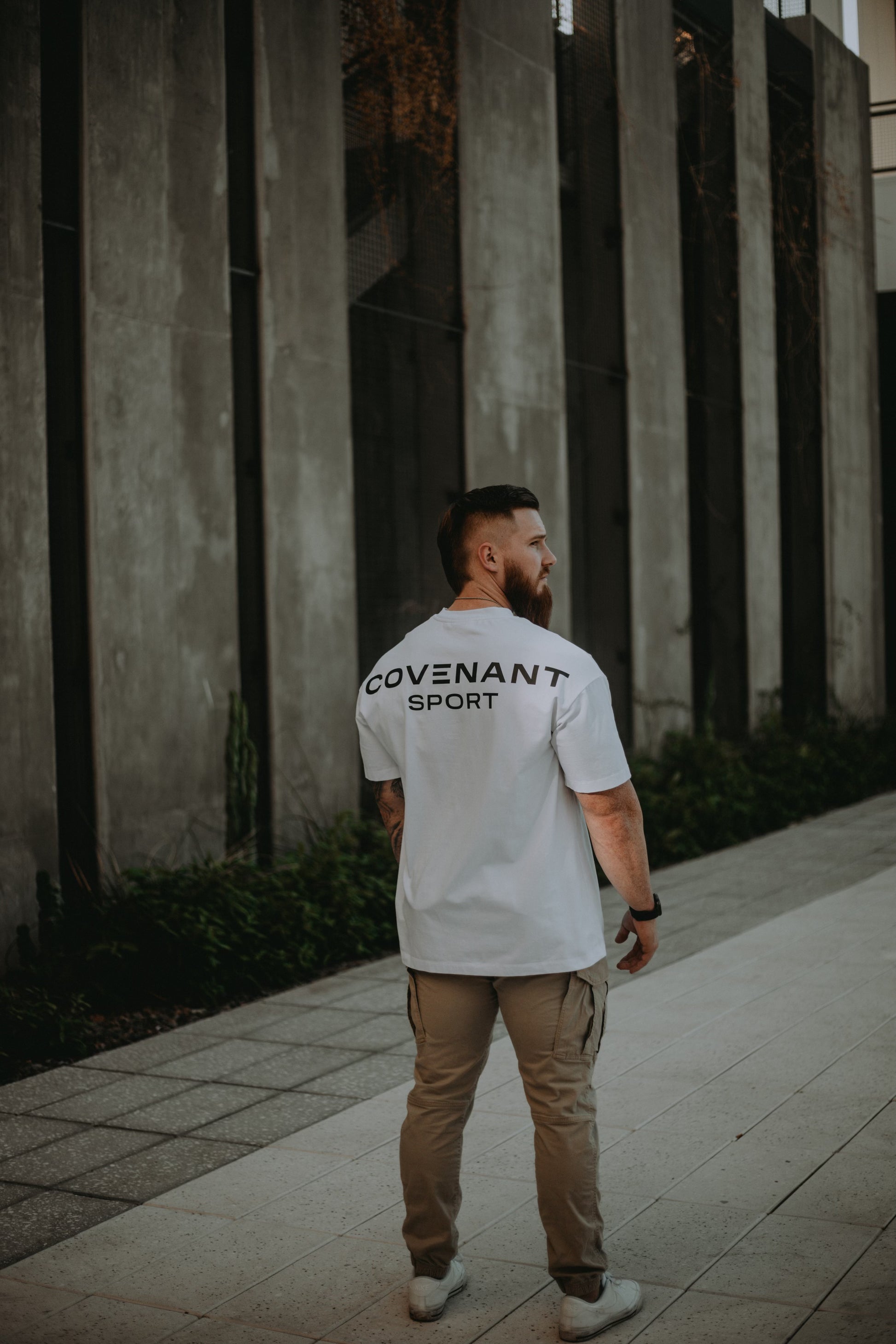 Model wearing the Brushed White Covenant Sport Tee in a street wear style. "Covenant Sport" is branded on the back of the tee from shoulder to shoulder in large, bold font. The tee is oversized and has a longer hem and has room in the back, chest, and shoulders. 